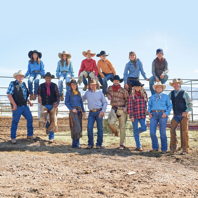 A group of cowboys and cowgirls bundled up in winter clothes and posed for the photo on and around a metal fence.